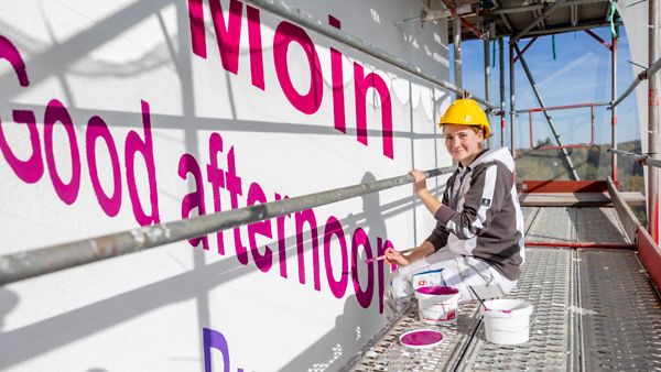Eine Auszubildende malt pinke Grußformeln an die Fassade des Gebäudes