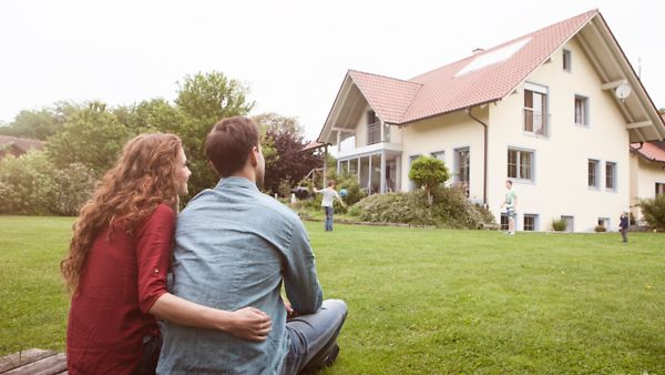 Eine Frau und ein Mann sitzen vor einem Einfamilienhaus, vor dem Kinder spielen