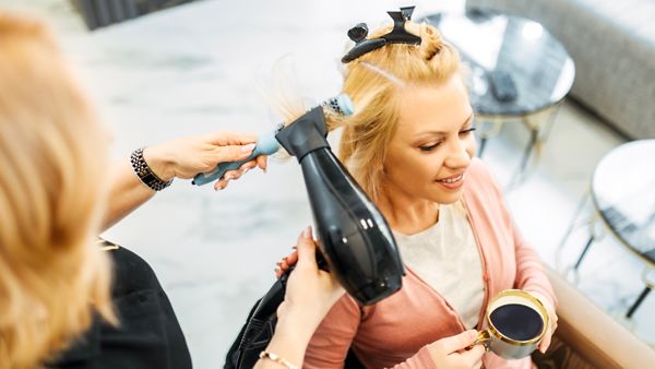 Eine Frau genießt ihren Kaffee, während eine Friseurin ihre Haare föhnt