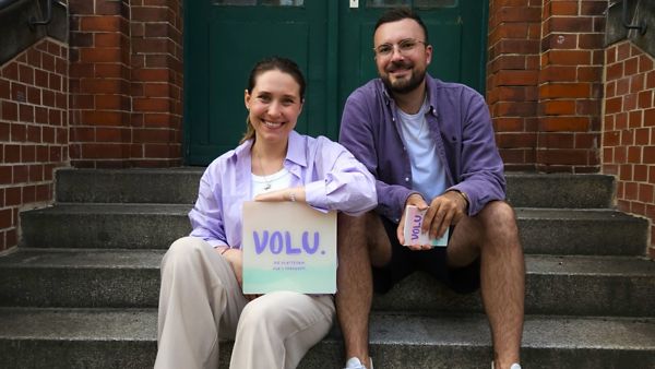 Die Gründerin und der Gründer von dem Startup Volu sitzen auf einer Treppe. Die Frau hält ein Schild mit dem Logo des Startups.