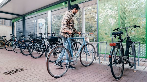 Mann mit einem blauen Fahrrad, welches er in eine Fahrradabstellanlage mit anderen Fahrrädern schiebt