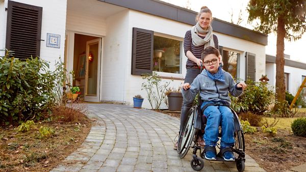 Eine junge Frau mit einem Kind im Rollstuhl, die vor einem barrierefreien und familiengerechten Haus stehen.